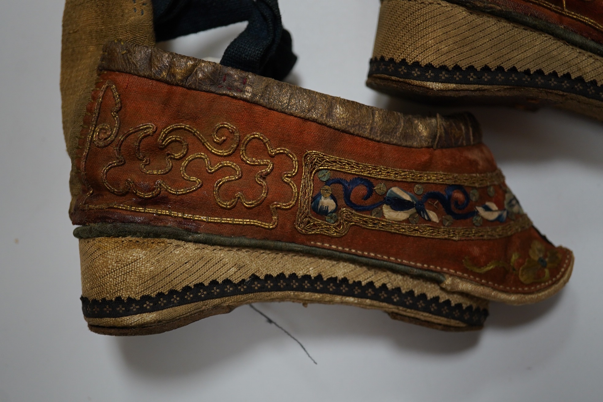 A pair of Chinese silk and gold thread embroidered ladies shoes, with leather binding around the top and gold brocade platform heels with fine blue braiding, 12cm long. Condition - good for age and use